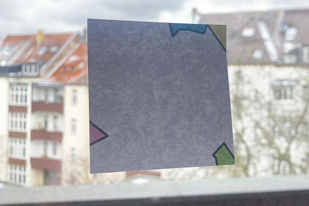 Ein Haftnotiz-Zettel, auch Post-it genannt, klebt an einer Fensterscheibe vor einer Häuserkulisse.