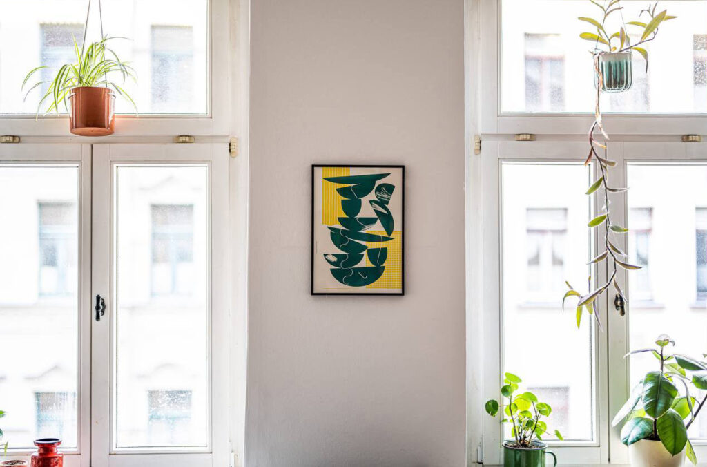 Schwarz gerahmtes Tellerstapel-Plakat in Grün-Gelb zwischen zwei Fenster in einer Altbau-Wohnung hängend