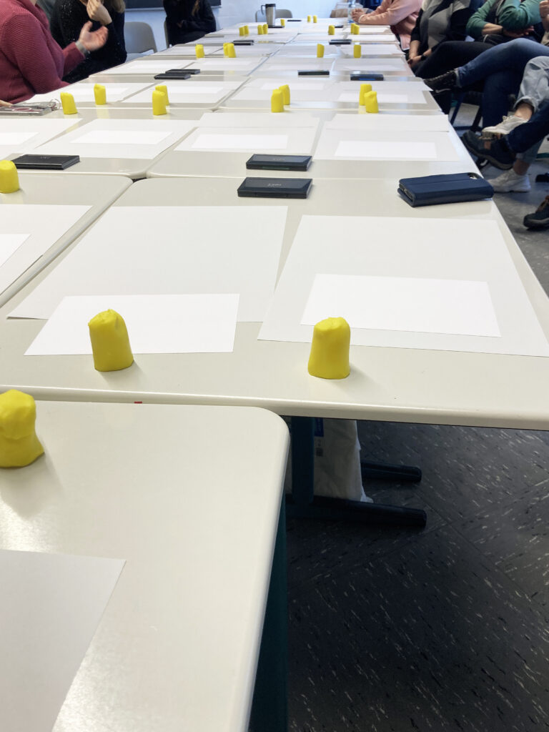 Knete, Papier und Stempelkissen liegen in dreißigfahcer Ausführung auf einem großen Tisch. Darum sitzen Lehrkräfte der Berufsschule.